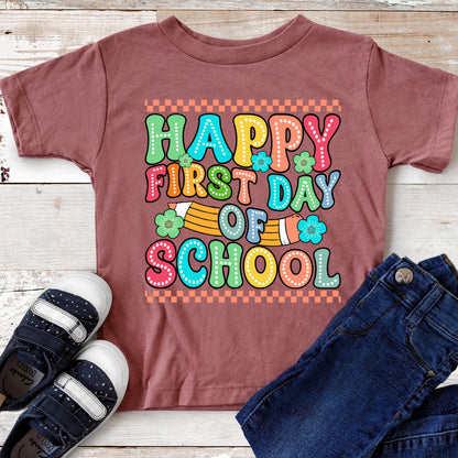 May include: A mauve short-sleeved t-shirt featuring a colorful graphic with the text HAPPY FIRST DAY OF SCHOOL in a retro bubble font. The design includes a yellow pencil illustration, small green flowers, and a checkered border pattern.