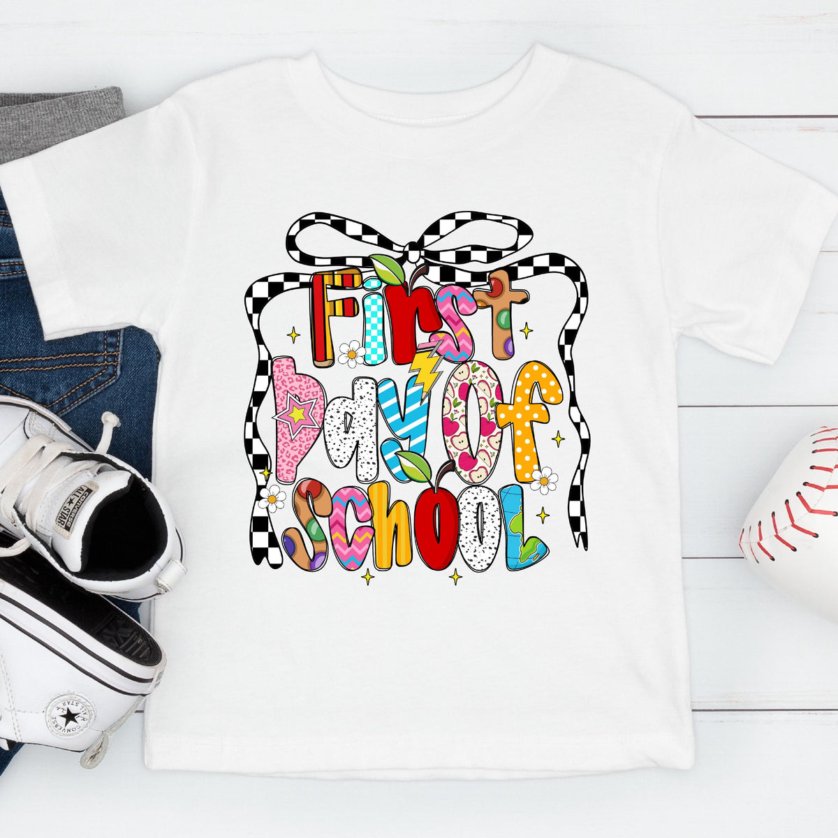 May include: White short-sleeved t-shirt with a colorful "First Day of School" graphic. The design features a mix of patterns and colors, including a checkered ribbon, flowers, and an apple. The shirt is displayed with a pair of jeans, sneakers, and a baseball.