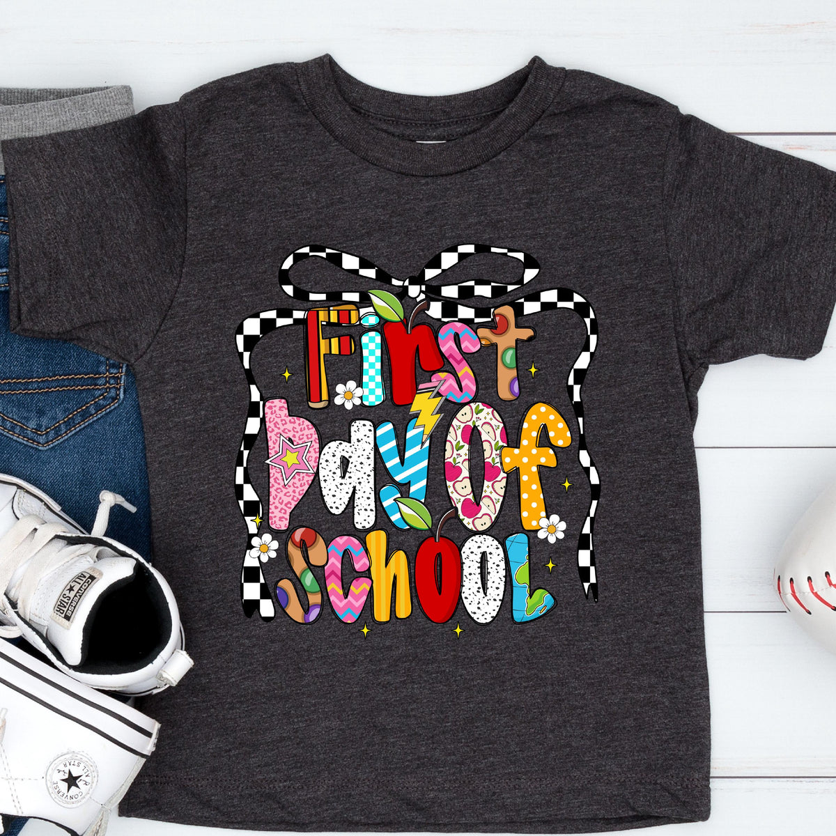 May include: Dark gray t-shirt with the text "First Day of School" in colorful, playful letters. The design includes a checkered bow and various decorative elements, perfect for back-to-school season.