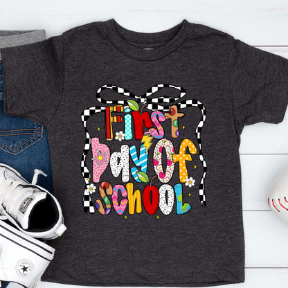 May include: Dark gray t-shirt with the text "First Day of School" in colorful, playful letters. The design includes a checkered bow and various decorative elements, perfect for back-to-school season.