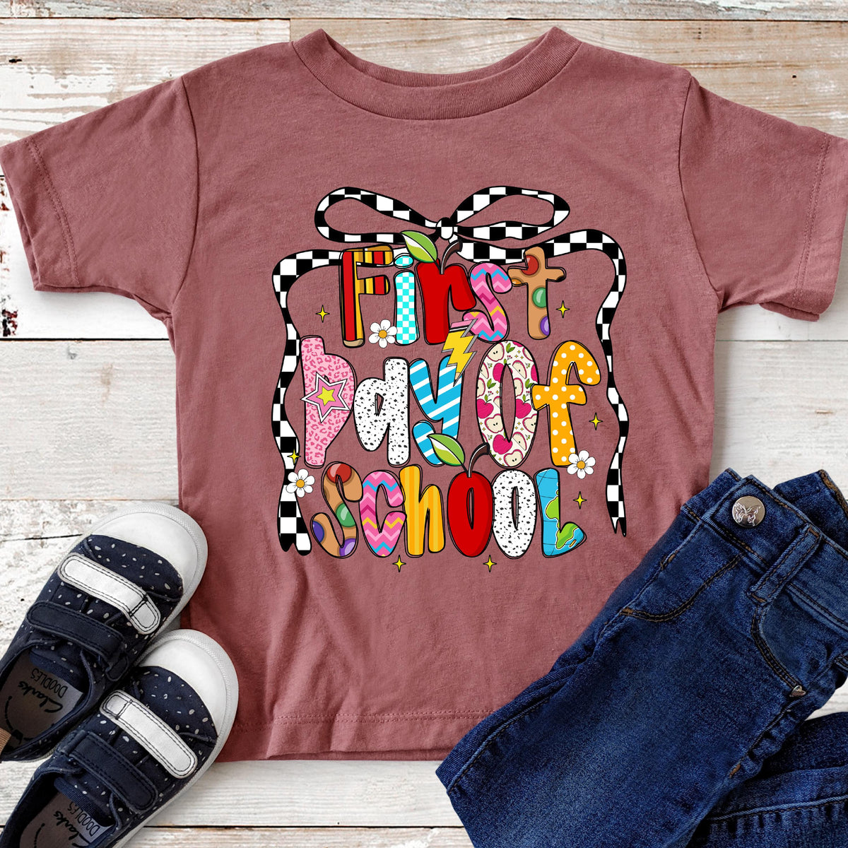 May include: A maroon short-sleeved t-shirt with the text "First Day Of School" in colorful, patterned letters. The shirt is paired with dark blue jeans and navy blue and white sneakers. A black and white checkered ribbon design frames the text.