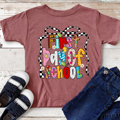 May include: A maroon short-sleeved t-shirt with the text "First Day Of School" in colorful, patterned letters. The shirt is paired with dark blue jeans and navy blue and white sneakers. A black and white checkered ribbon design frames the text.