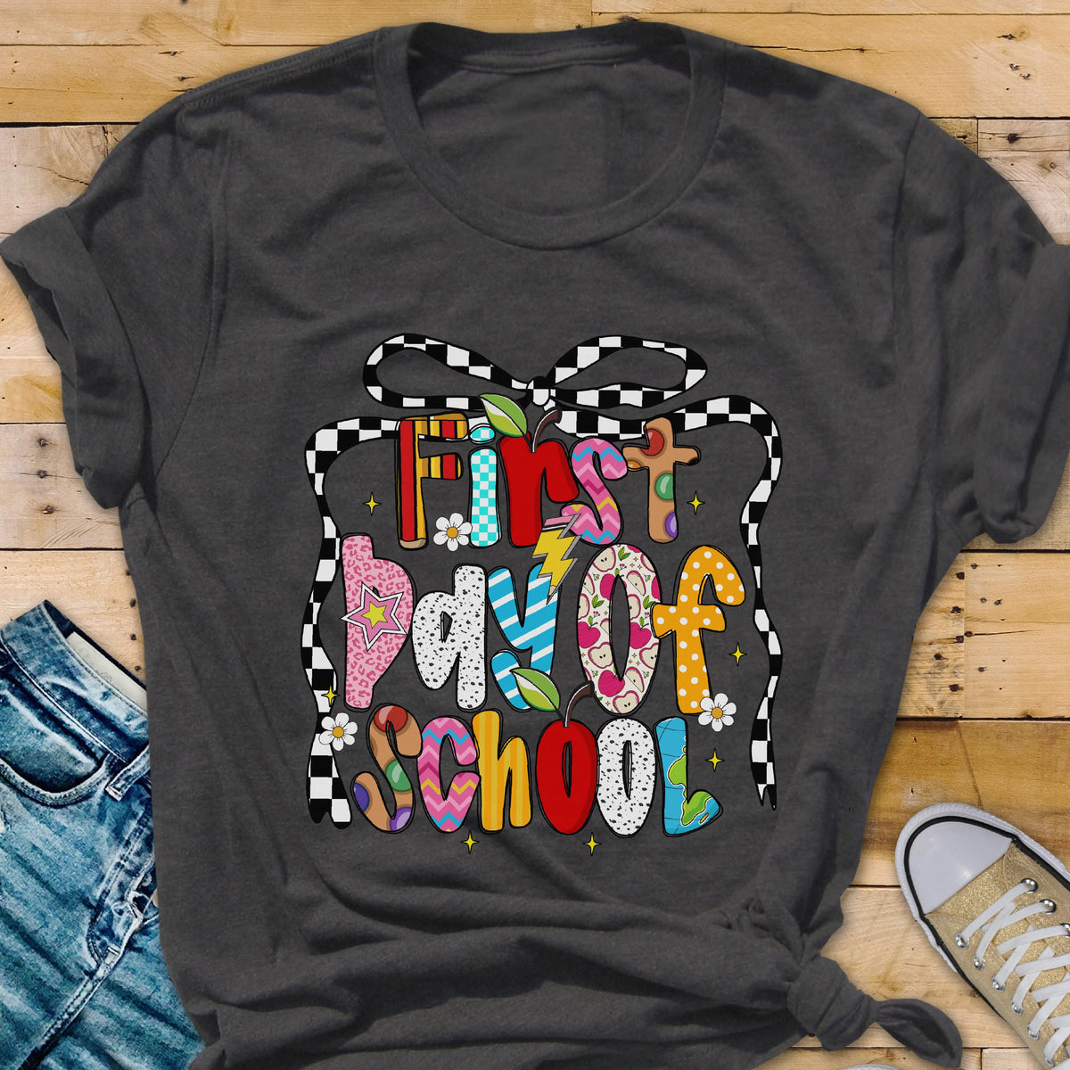 May include: A dark gray t-shirt featuring a colorful graphic with the text "First day of SCHOOL" in various patterns and colors. The design is framed by a black and white checkered ribbon outline, accented with small flowers and a lightning bolt.