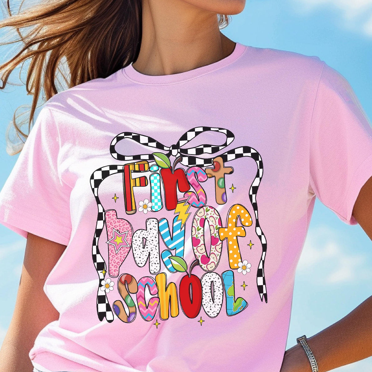May include: A light pink t-shirt featuring a graphic print with the text First Day of School. The design includes a black and white checkered bow, colorful letters with various patterns like polka dots and stripes, and small apple icons.