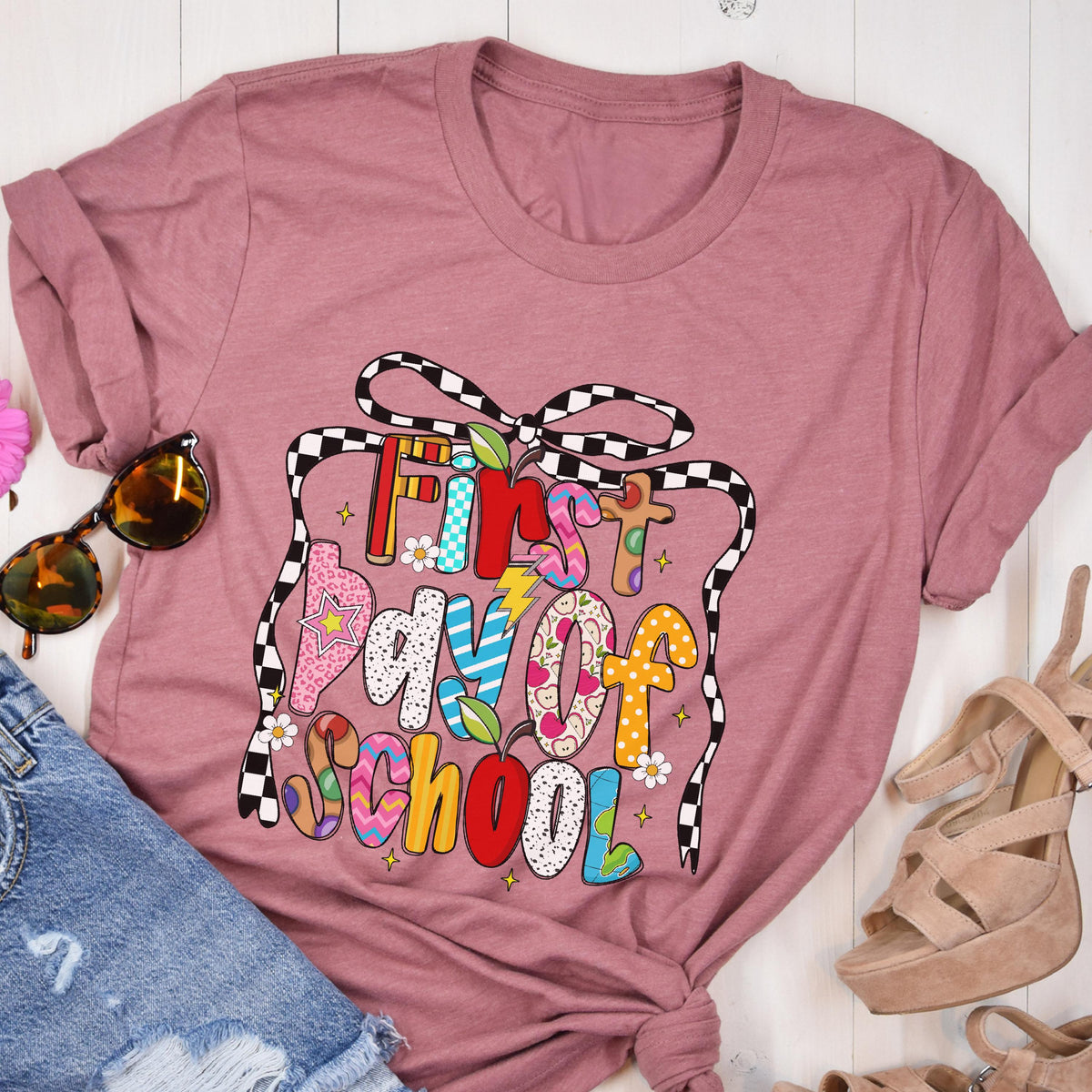 May include: A mauve short-sleeved t-shirt featuring a graphic print of a black and white checkered bow framing the text First Day of School. The letters are filled with various colorful patterns including stripes, polka dots, and floral designs.