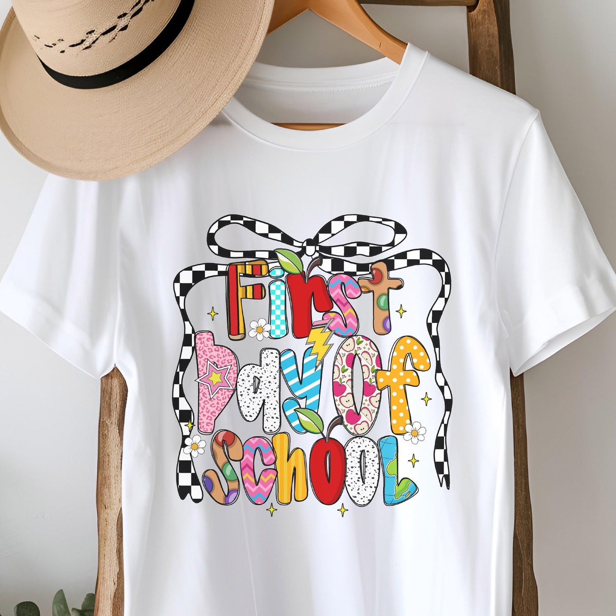 May include: A white t-shirt featuring a graphic print with the text First Day of School in colorful, patterned bubble letters. The design includes a black and white checkered bow, small yellow stars, and white flowers, all centered on the chest.