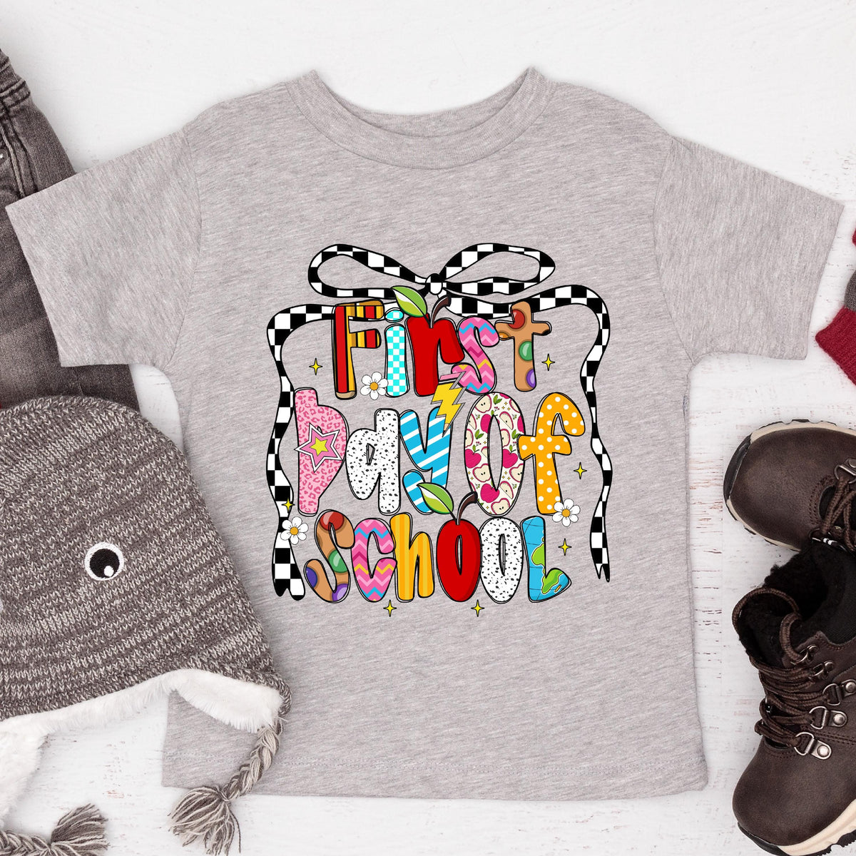 May include: A light gray toddler t-shirt with a colorful graphic that reads "First Day of School." The text is in various colors and patterns, including flowers, stars, and a checkered ribbon design. The shirt is surrounded by a gray hat and brown boots.