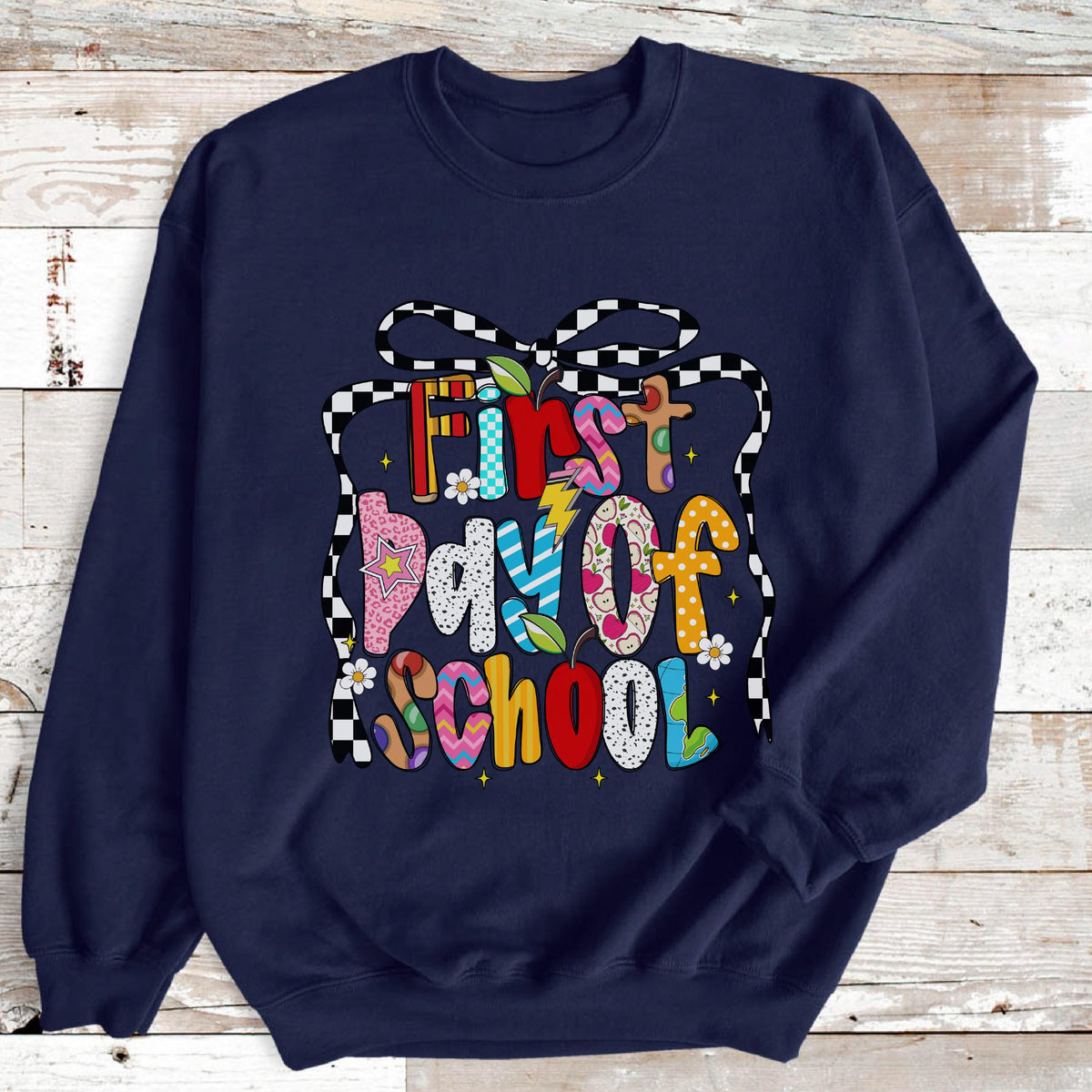 May include: A navy blue crewneck sweatshirt featuring a colorful graphic print that reads First Day of School. The text is surrounded by a black and white checkered bow design, with various patterns including stars, dots, and stripes in bright colors.