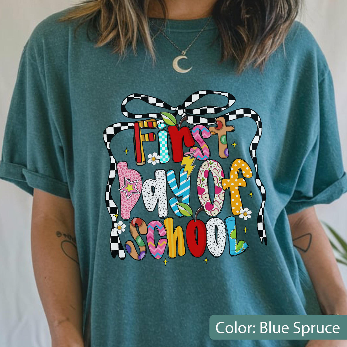 May include: Blue spruce t-shirt with a colorful graphic that reads "First Day of School." The text is designed with various patterns, including flowers, lightning bolts, and polka dots. A black and white checkered ribbon frames the text.