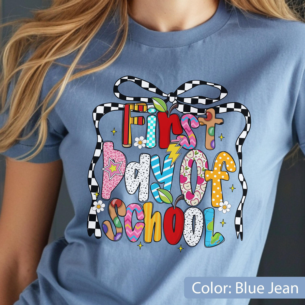 May include: Blue jean colored t-shirt with a colorful graphic that says "First Day of School." The text is designed with various patterns and colors, including red, pink, and blue. A black and white checkered ribbon frames the text.