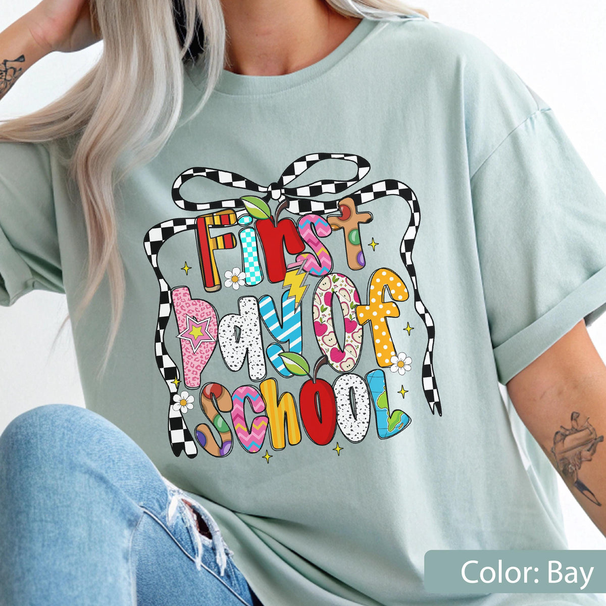 May include: A light blue t-shirt with a colorful graphic that says "First Day of School." The text is in various colors and patterns, including red, pink, yellow, and blue. The shirt has a relaxed fit and short sleeves.