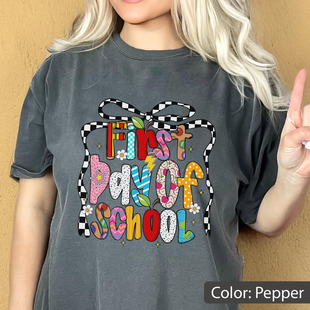 May include: Pepper-colored t-shirt with a colorful "First Day Of School" graphic. The design features a gift box outline with a black and white checkered ribbon. The text is in various patterns and colors, including pink, blue, and yellow.