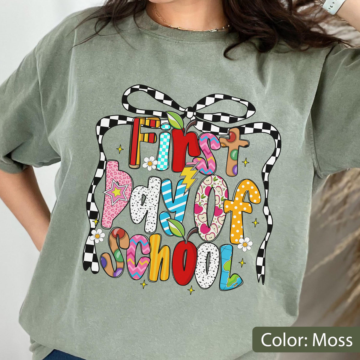 May include: Moss green t-shirt with a colorful graphic that reads "First Day of School." The text is designed with various patterns and colors, including polka dots, stripes, and floral prints. A black and white checkered bow frames the text.