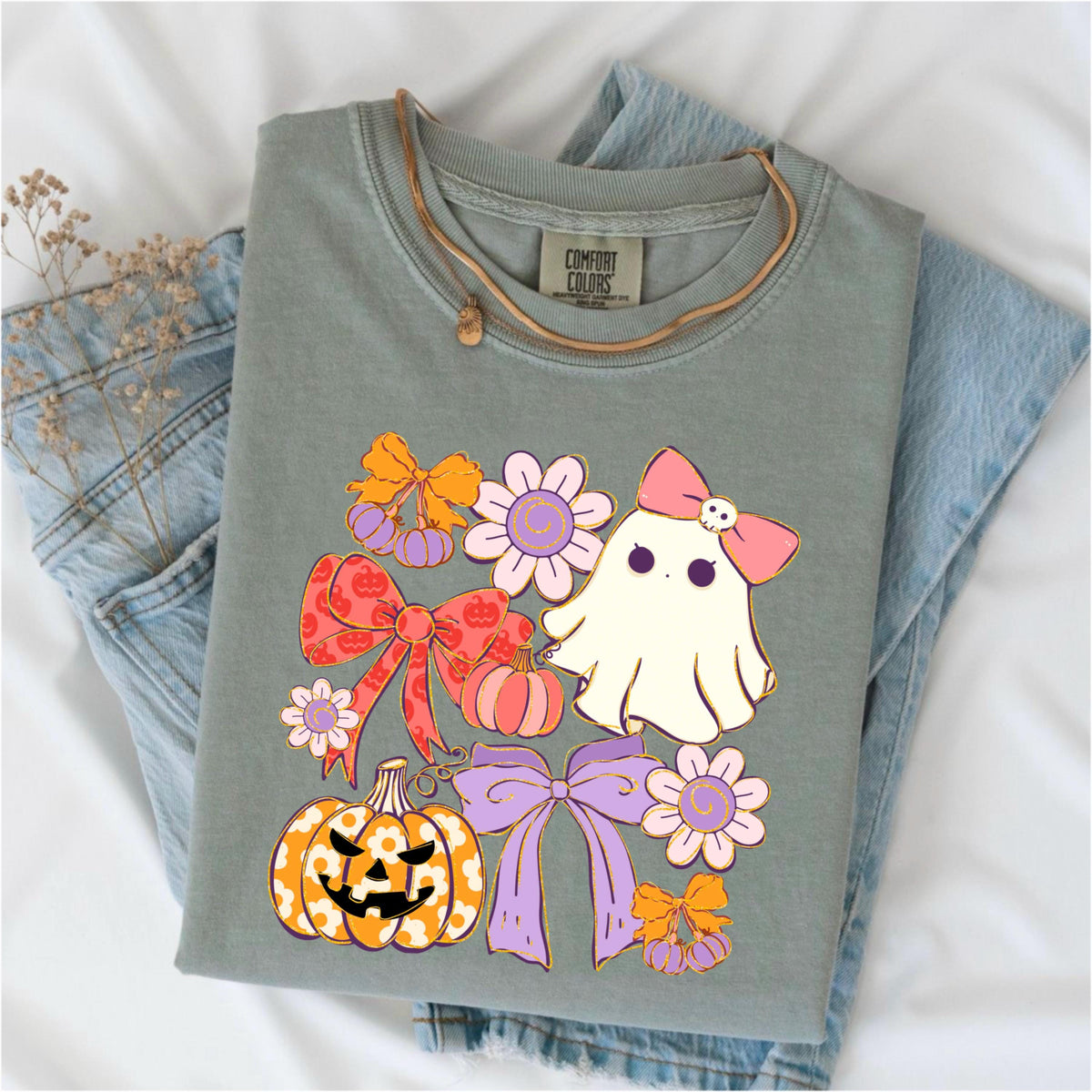 May include: Sage green Comfort Colors t-shirt with a Halloween-themed graphic. The design features a cute ghost with a pink bow, pumpkins, bows, and flowers in shades of orange, pink, and purple. The shirt is folded on top of blue jeans.
