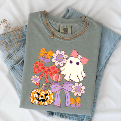 May include: Sage green Comfort Colors t-shirt with a Halloween-themed graphic. The design features a cute ghost with a pink bow, pumpkins, bows, and flowers in shades of orange, pink, and purple. The shirt is folded on top of blue jeans.