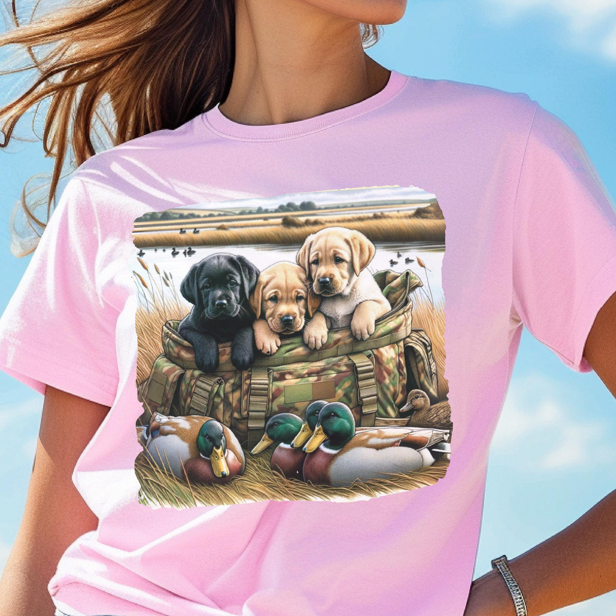 May include: A light pink t-shirt featuring a graphic of three Labrador puppies in a camouflage bag, with ducks in the foreground. The puppies are black, tan, and yellow. The background shows a marshy landscape.