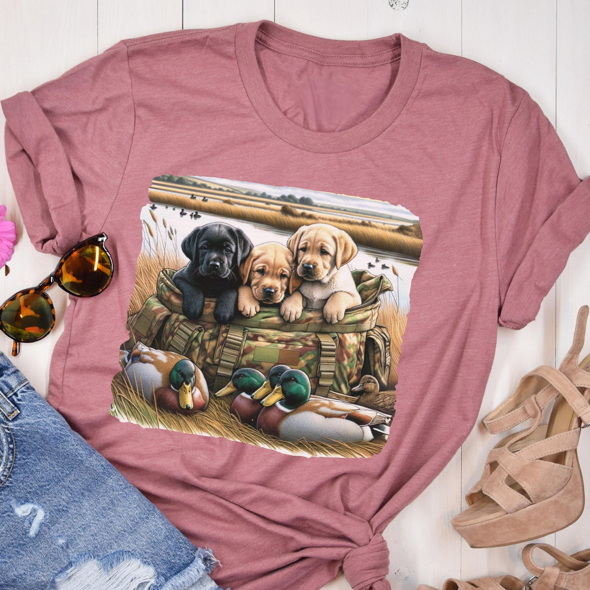 May include: A mauve-colored t-shirt featuring a graphic of three Labrador Retriever puppies in a camouflage bag, with ducks in the foreground and a marsh scene in the background. The puppies are black, tan, and yellow.