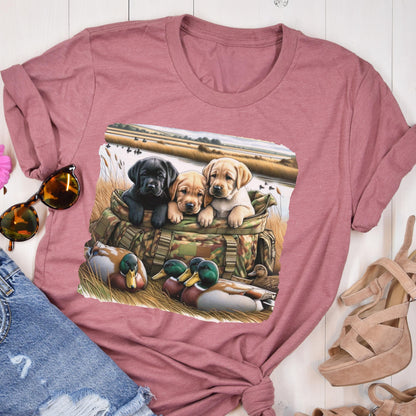 May include: A mauve-colored t-shirt featuring a graphic of three Labrador Retriever puppies in a camouflage bag, with ducks in the foreground and a marsh scene in the background. The puppies are black, tan, and yellow.