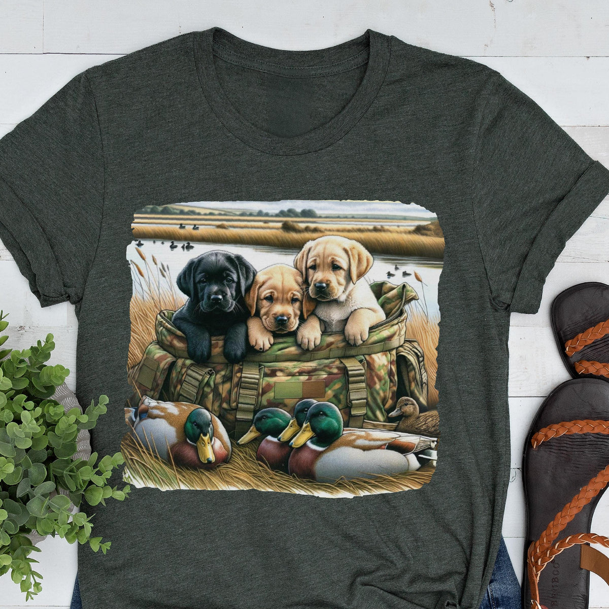 May include: Dark gray t-shirt featuring a graphic of three Labrador puppies in a camouflage bag, with ducks in the foreground. The puppies are black, tan, and yellow. The background shows a marsh scene.