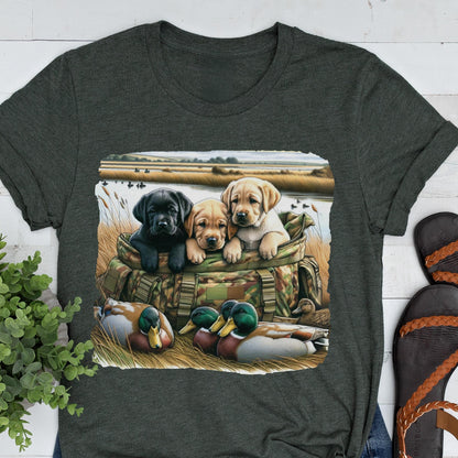 May include: Dark gray t-shirt featuring a graphic of three Labrador puppies in a camouflage bag, with ducks in the foreground. The puppies are black, tan, and yellow. The background shows a marsh scene.
