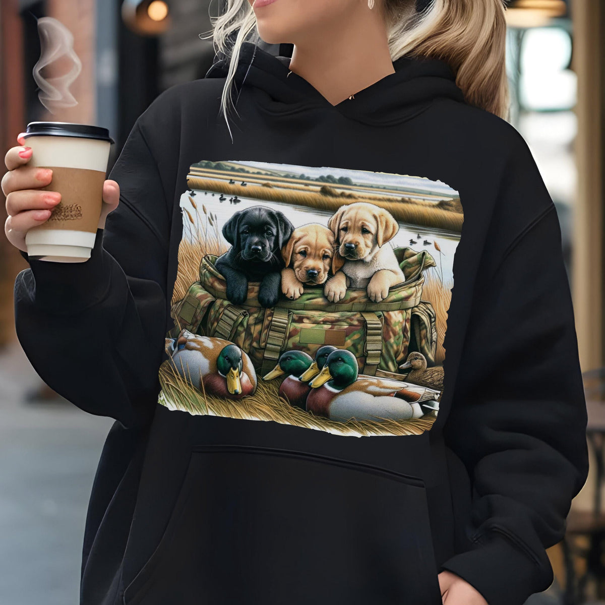 May include: A black hooded sweatshirt featuring a graphic print of three Labrador retriever puppies sitting inside a camouflage bag, with two mallard ducks resting in the foreground grass near a body of water and a marshy landscape.