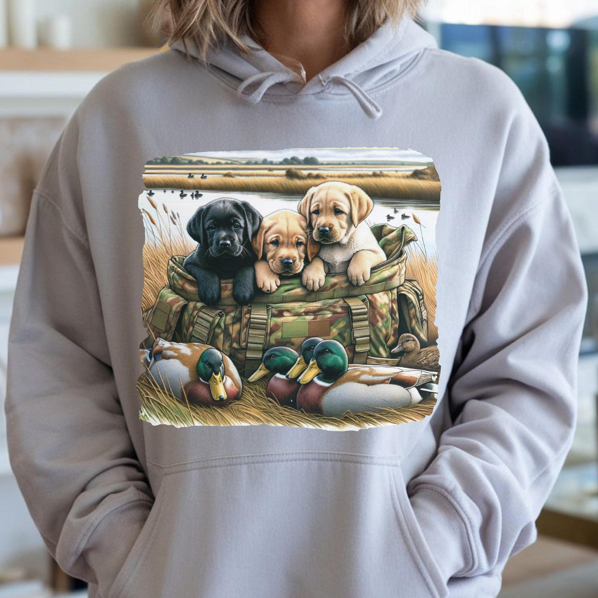 May include: A light gray hooded sweatshirt featuring a graphic print of three Labrador retriever puppies sitting inside a camouflage hunting bag, with two mallard ducks resting in the tall grass in the foreground of the outdoor scene.