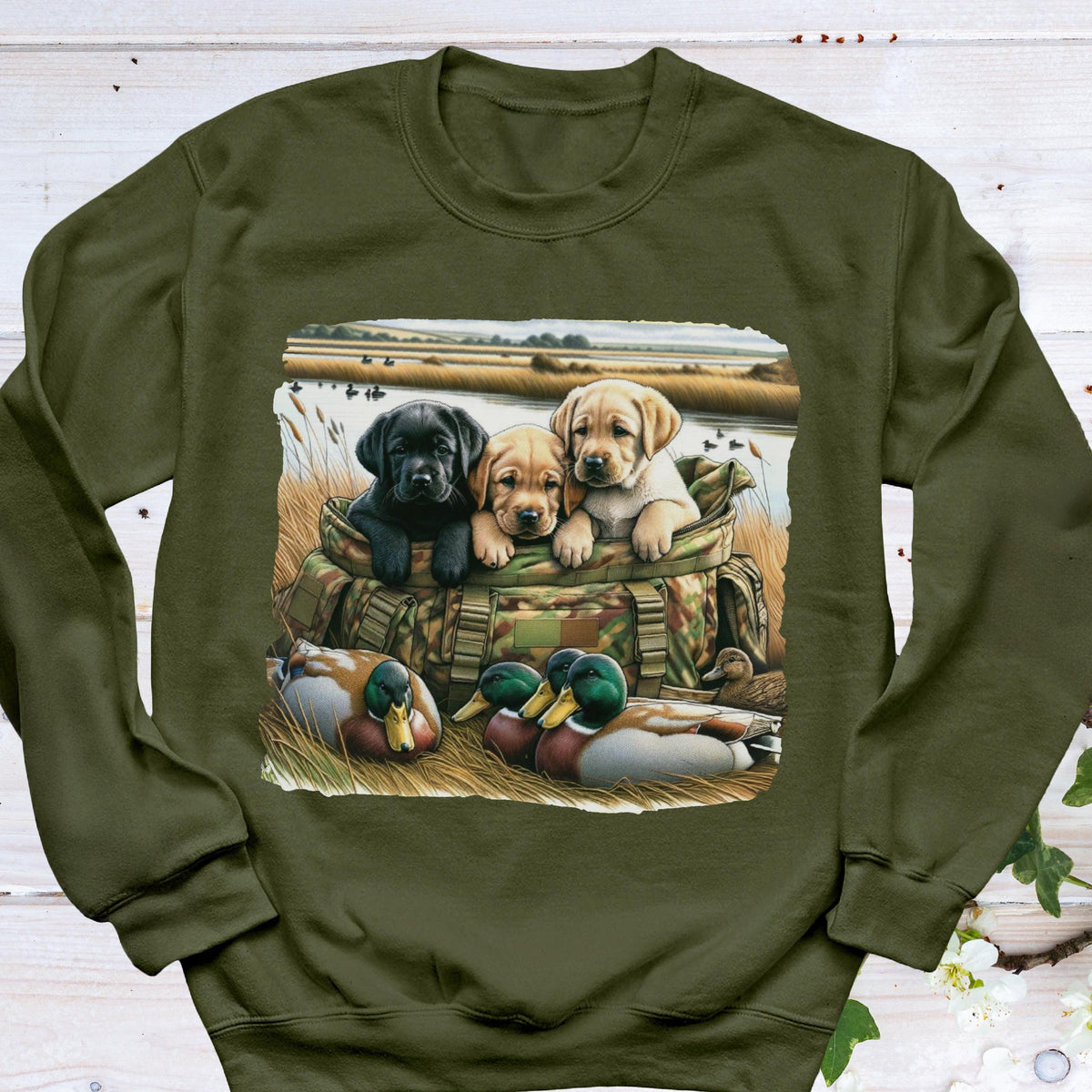 May include: An olive green crewneck sweatshirt featuring a graphic print of three puppies, one black and two yellow, sitting inside a camouflage hunting bag. In the foreground, two mallard ducks rest in the grass near a body of water.