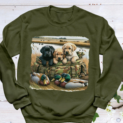 May include: An olive green crewneck sweatshirt featuring a graphic print of three puppies, one black and two yellow, sitting inside a camouflage hunting bag. In the foreground, two mallard ducks rest in the grass near a body of water.