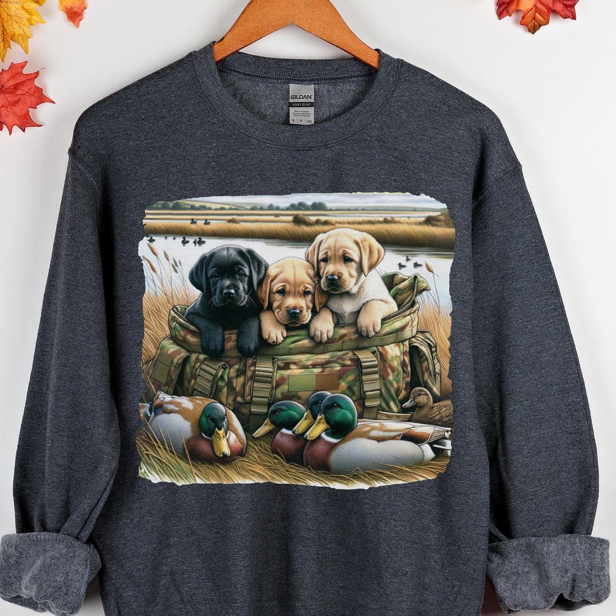 May include: A dark gray crewneck sweatshirt featuring a graphic print of three Labrador retriever puppies sitting inside a camouflage hunting bag, with two mallard ducks resting in the tall grass in the foreground near a body of water.