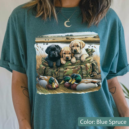 May include: A blue spruce t-shirt featuring a graphic print of three Labrador retriever puppies sitting inside a camouflage duffel bag, with two mallard ducks resting in the tall grass in the foreground of a marshy landscape scene.