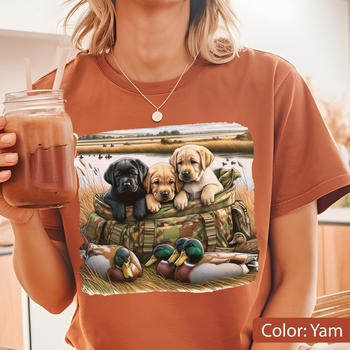 May include: A yam-colored t-shirt featuring a graphic print of three Labrador retriever puppies, one black and two yellow, sitting inside a camouflage hunting bag. In the foreground, two mallard ducks rest in the grass near a body of water.
