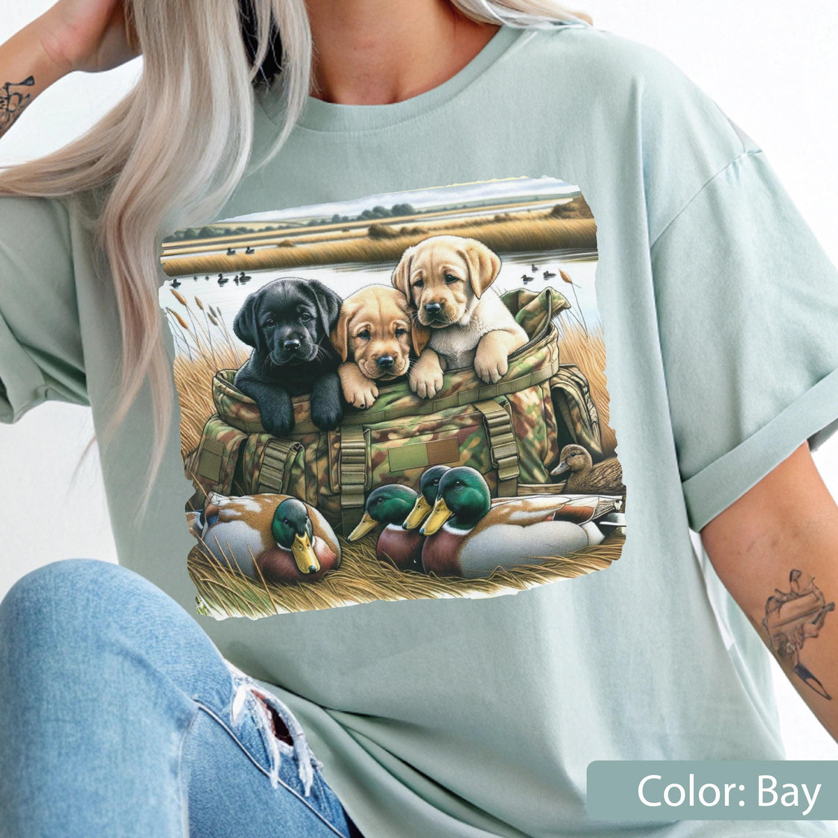May include: A light sage green t-shirt featuring a graphic print of three Labrador retriever puppies, one black and two yellow, sitting inside a camouflage hunting bag. Two mallard ducks are resting in the foreground grass near the bag.