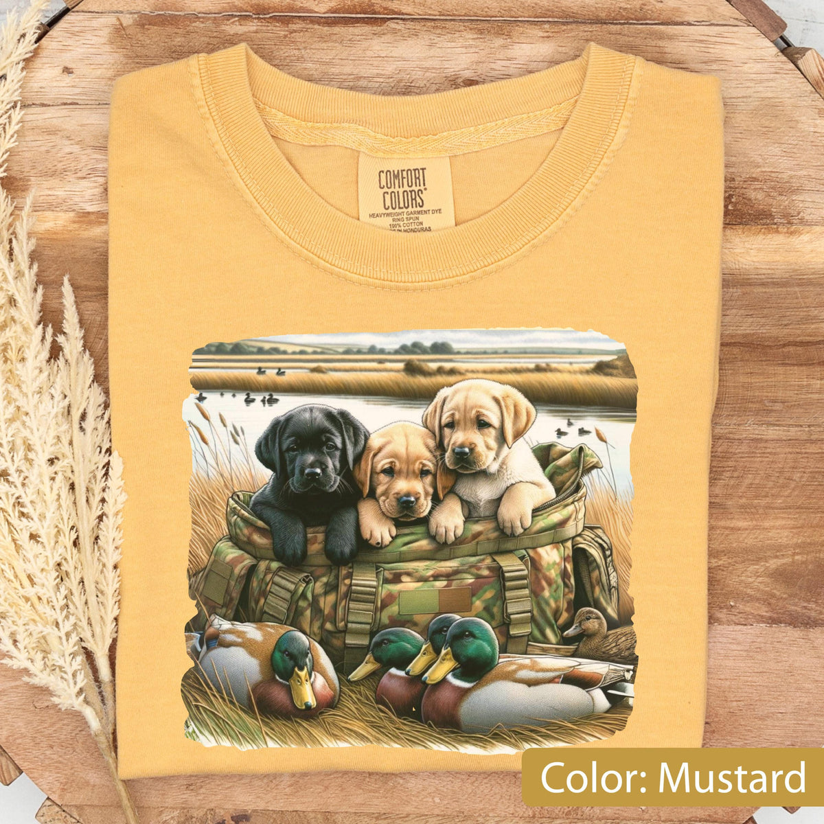 May include: A mustard yellow t-shirt featuring a graphic print of three puppies, one black and two yellow, sitting inside a camouflage hunting bag. In the foreground, two mallard ducks rest in tall grass near a body of water with a field background.