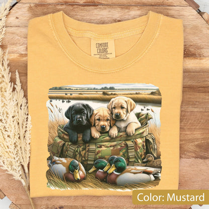 May include: A mustard yellow t-shirt featuring a graphic print of three puppies, one black and two yellow, sitting inside a camouflage hunting bag. In the foreground, two mallard ducks rest in tall grass near a body of water with a field background.