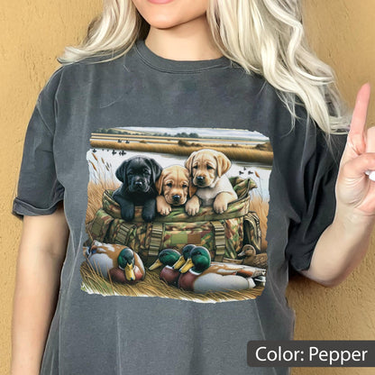May include: A pepper-colored t-shirt featuring a graphic print of three Labrador retriever puppies sitting inside a camouflage hunting bag, with two mallard ducks resting in the foreground grass near a body of water and a field.