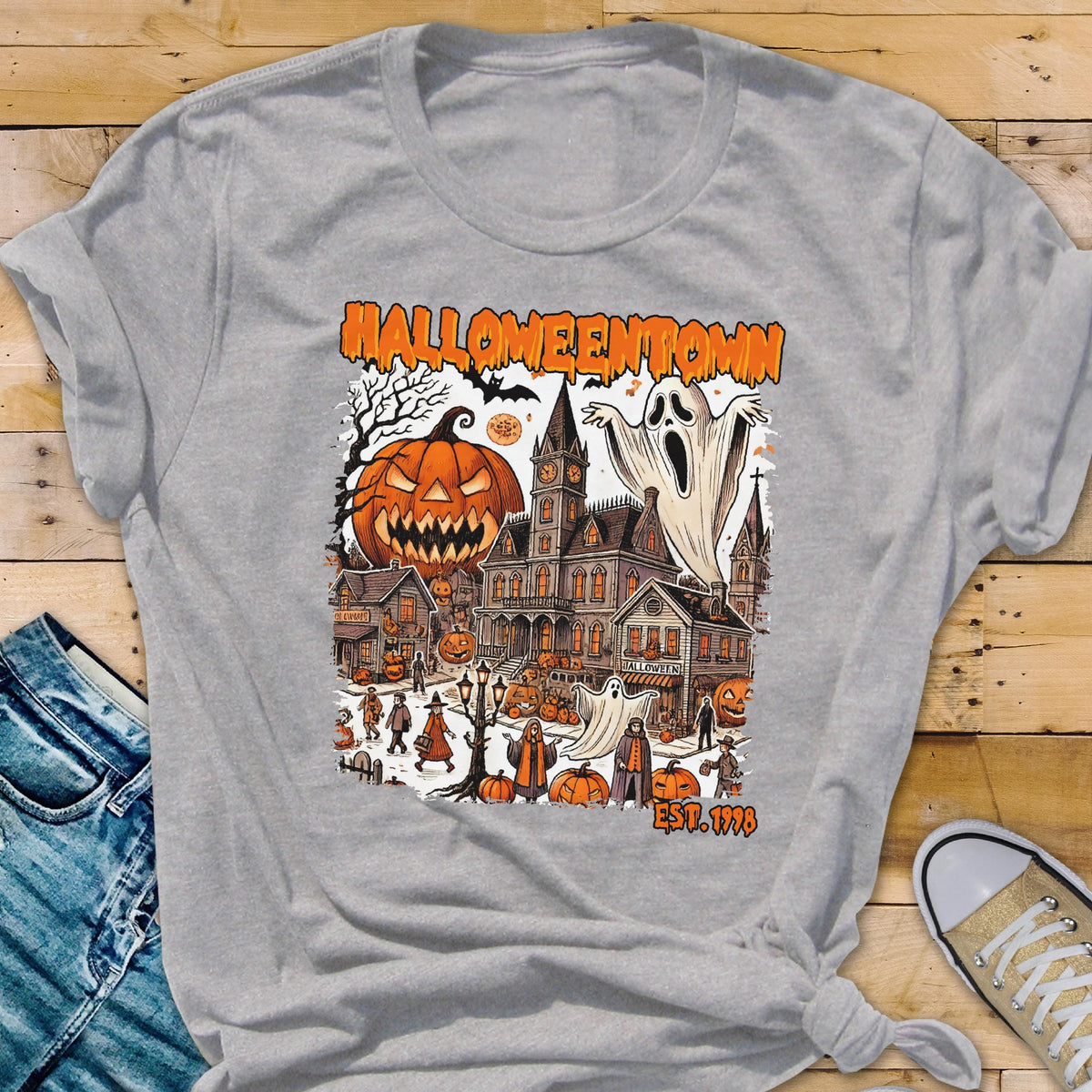May include: A gray t-shirt featuring a Halloween-themed graphic. The design includes a jack-o'-lantern, a haunted house, ghosts, and the text "Halloweentown EST. 1998". The graphic is in orange, black, and white.