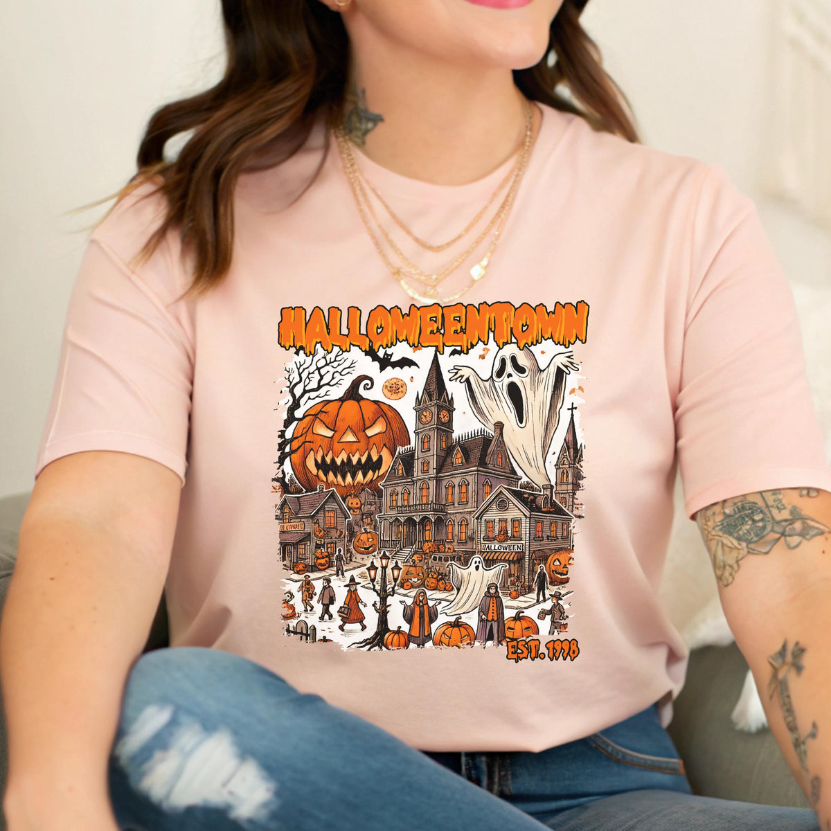 May include: Pink t-shirt featuring a Halloween-themed graphic. The design includes a jack-o'-lantern, a haunted house, ghosts, and the text "Halloweentown EST. 1998". The graphic is in shades of orange, black, and white.