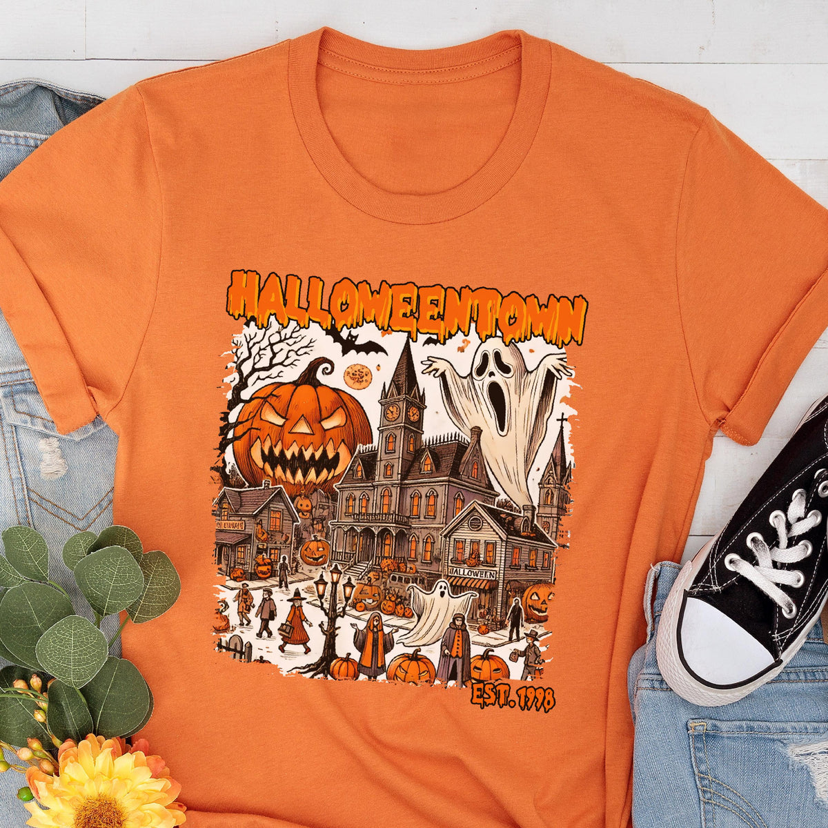 May include: Orange t-shirt featuring a Halloween-themed graphic. The design includes a jack-o'-lantern, a haunted house, ghosts, and the text "Halloweentown" and "Est. 1998". The shirt is paired with blue jeans and a black sneaker.