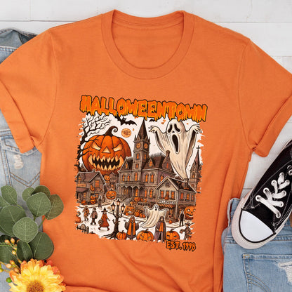 May include: Orange t-shirt featuring a Halloween-themed graphic. The design includes a jack-o'-lantern, a haunted house, ghosts, and the text "Halloweentown" and "Est. 1998". The shirt is paired with blue jeans and a black sneaker.