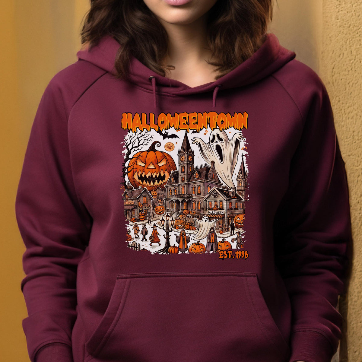 May include: A maroon hoodie featuring a graphic print with the text HALLOWEENTOWN EST. 1998. The illustration depicts a spooky town scene with a large jack-o'-lantern, a haunted clock tower, ghosts, and various figures in a dark, festive setting.