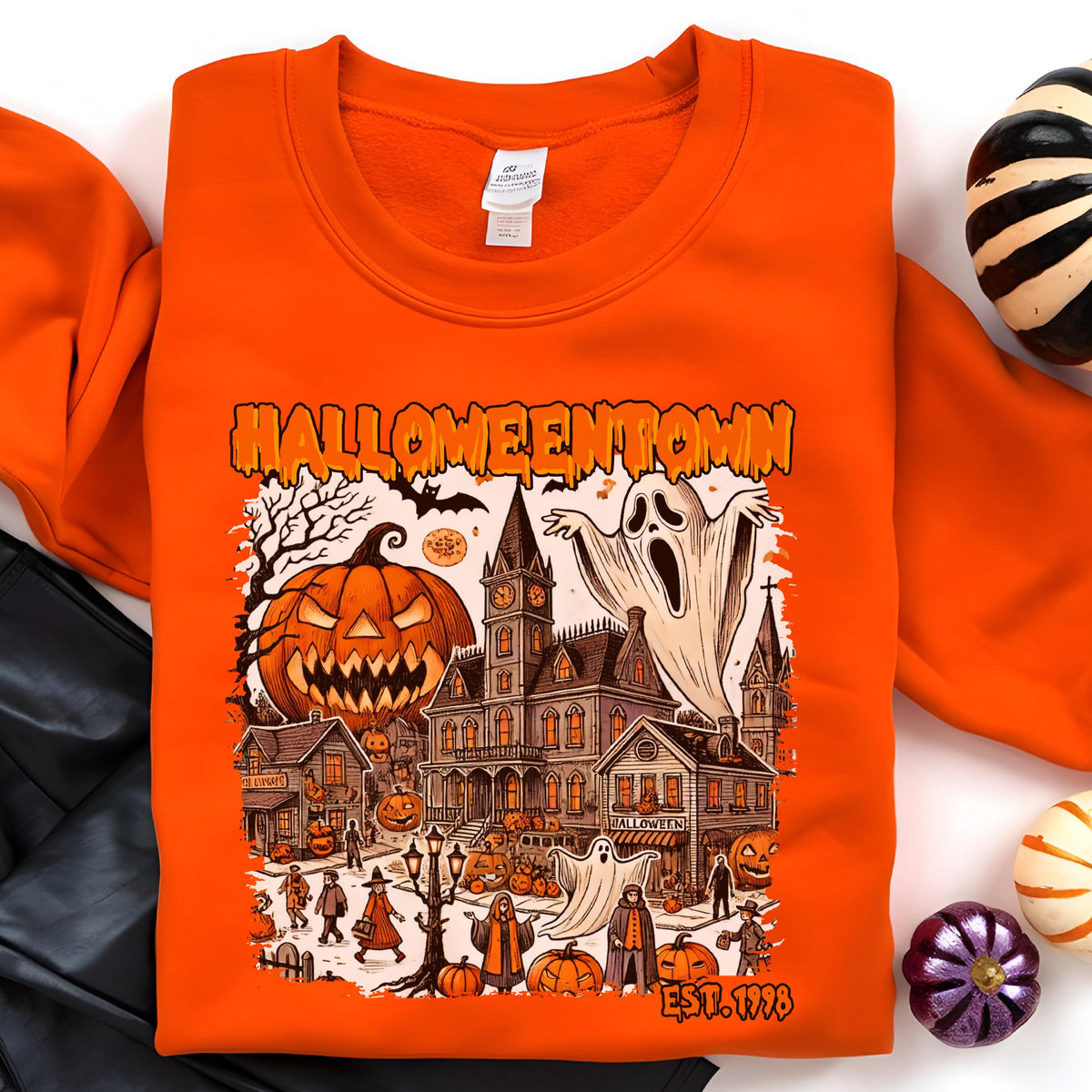 May include: An orange crewneck sweatshirt featuring a graphic print with the text HALLOWEENTOWN and EST. 1998. The illustration depicts a spooky town scene with a large jack-o'-lantern, a haunted mansion, ghosts, and various figures in costumes.