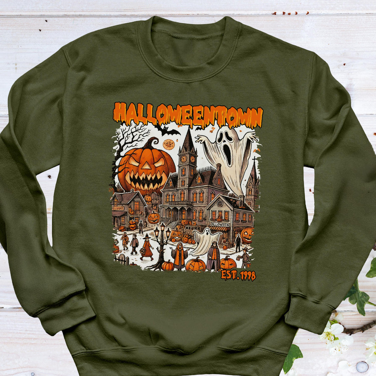 May include: An olive green crewneck sweatshirt featuring a graphic print of a spooky town scene with a large jack-o'-lantern, a ghost, and a gothic building. The text HALLOWEENTOWN and EST. 1998 are printed in orange above and below the illustration.