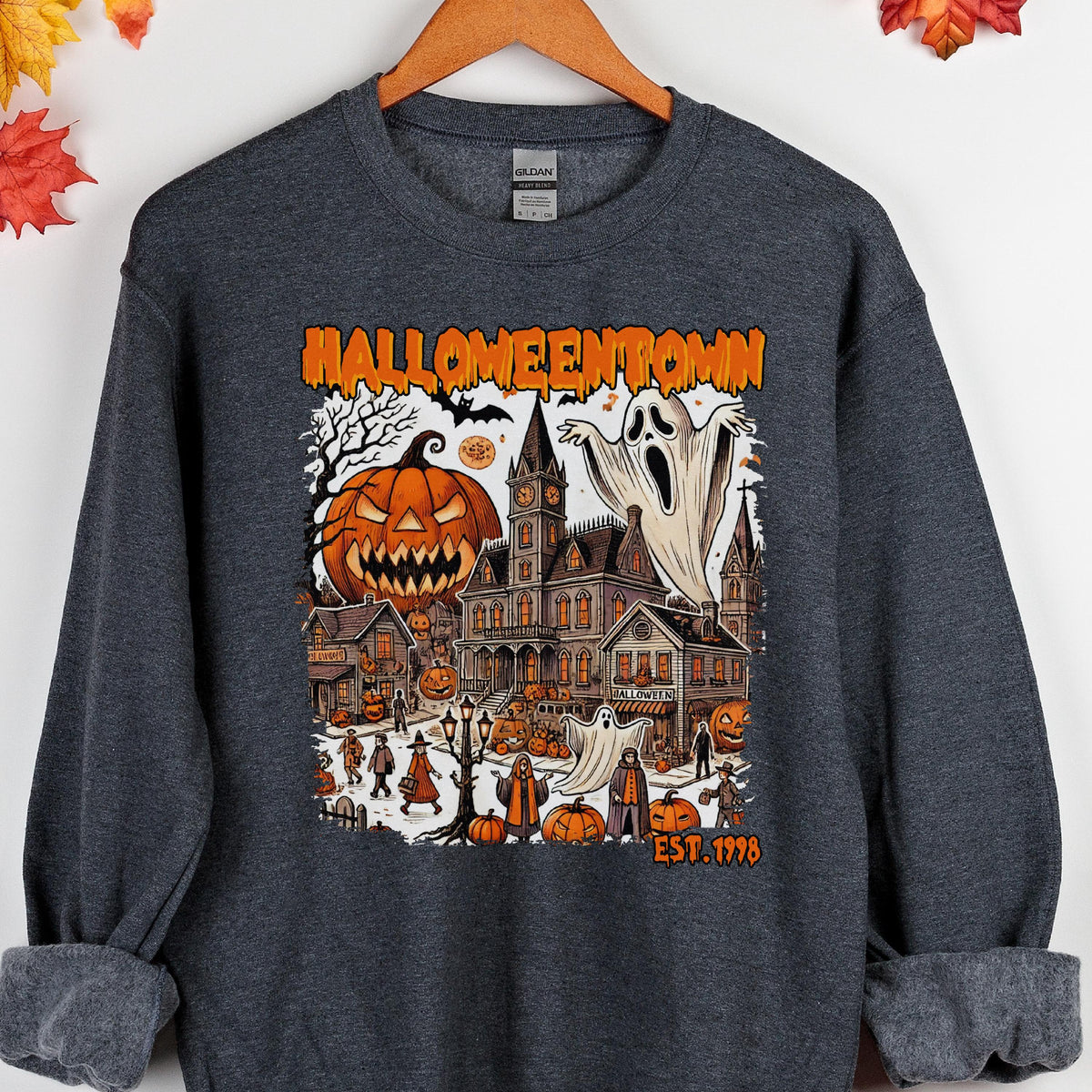May include: A dark heather gray crewneck sweatshirt featuring a graphic print with the text HALLOWEENTOWN EST. 1998. The design depicts a spooky town scene with a large jack-o'-lantern, a tall clock tower, ghosts, and figures in costumes.