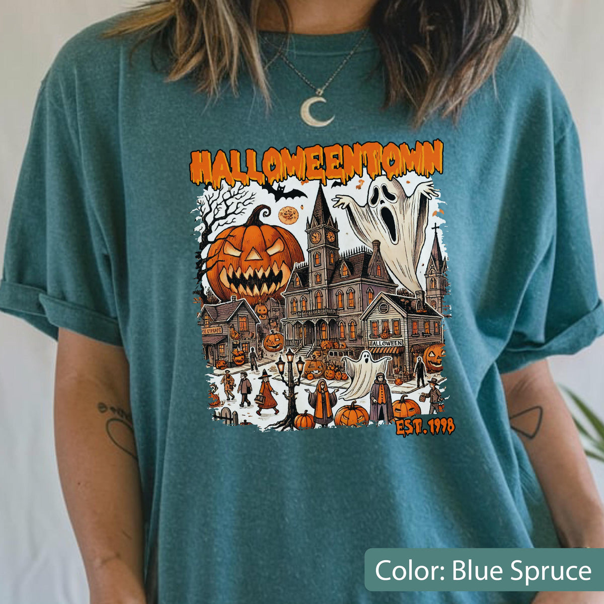 May include: A blue-spruce t-shirt featuring a graphic print with the text HALLOWEENTOWN EST. 1998. The design includes a large jack-o'-lantern, a haunted house, ghosts, and various figures in a town setting with orange and black color accents.