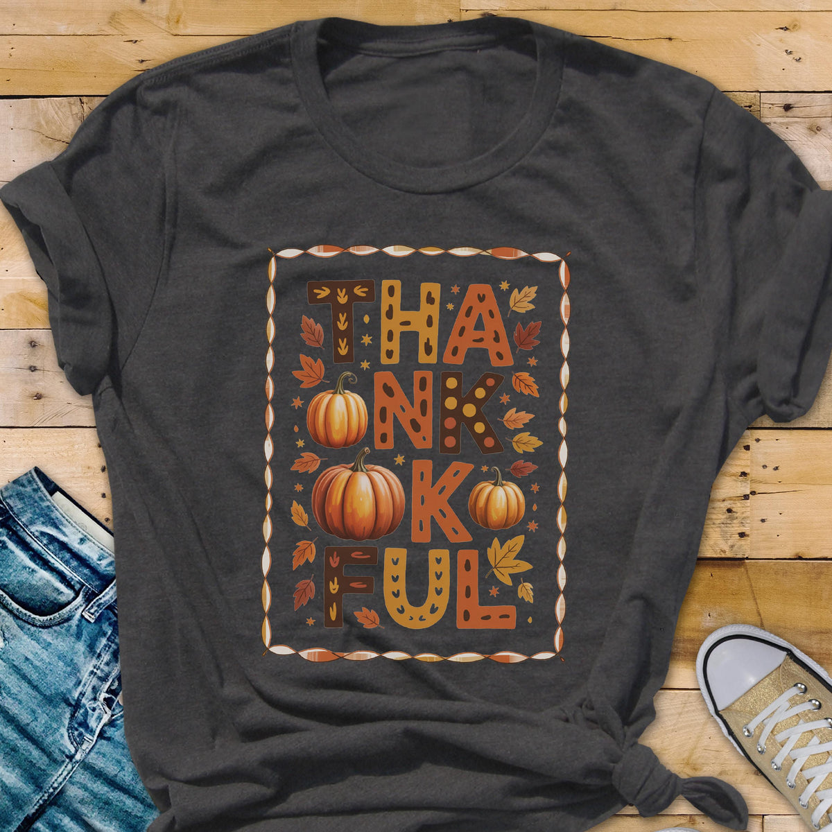 May include: A dark gray short-sleeve t-shirt featuring a rectangular graphic with the word THANKFUL in large block letters. The design includes orange pumpkins and autumn leaves in shades of yellow, orange, and brown, all enclosed in a decorative border.