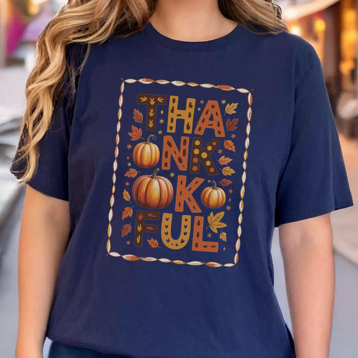 May include: A navy blue t-shirt featuring a rectangular graphic with the word THANKFUL in large, patterned letters. The design includes three orange pumpkins and various autumn leaves in shades of orange, yellow, and brown, all enclosed in a border.