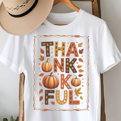May include: A white short-sleeved t-shirt featuring a rectangular border containing the text THANKFUL in brown and orange patterned letters. The design includes three orange pumpkins and scattered autumn leaves in shades of yellow and brown.