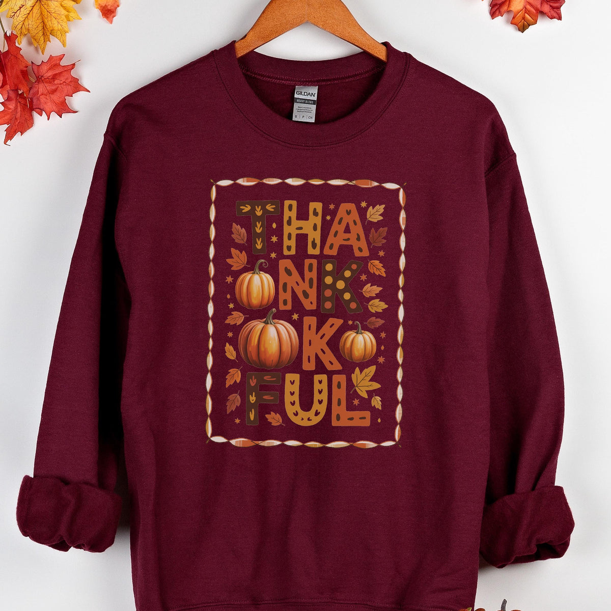 May include: A maroon sweatshirt with a fall-themed graphic. The graphic features the word "THANKFUL" in orange and brown letters, with pumpkins and leaves. The sweatshirt has a crew neck and long sleeves.