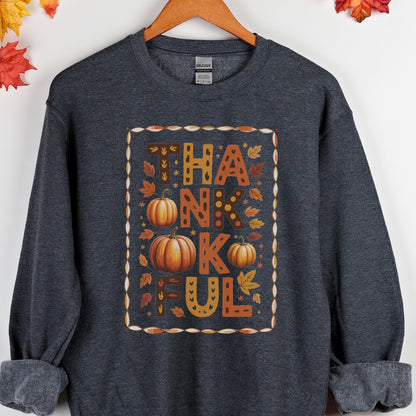 May include: Dark gray sweatshirt with a fall-themed design. The design features the word "THANKFUL" in orange and brown, with pumpkins and leaves. The sweatshirt is hanging on a wooden hanger.