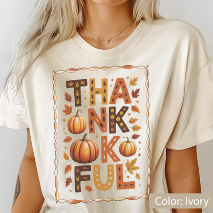 May include: Ivory-colored t-shirt with the word "THANKFUL" in a fall-themed design. The design includes pumpkins and autumn leaves in shades of orange, brown, and yellow. The shirt has a crew neck and short sleeves.